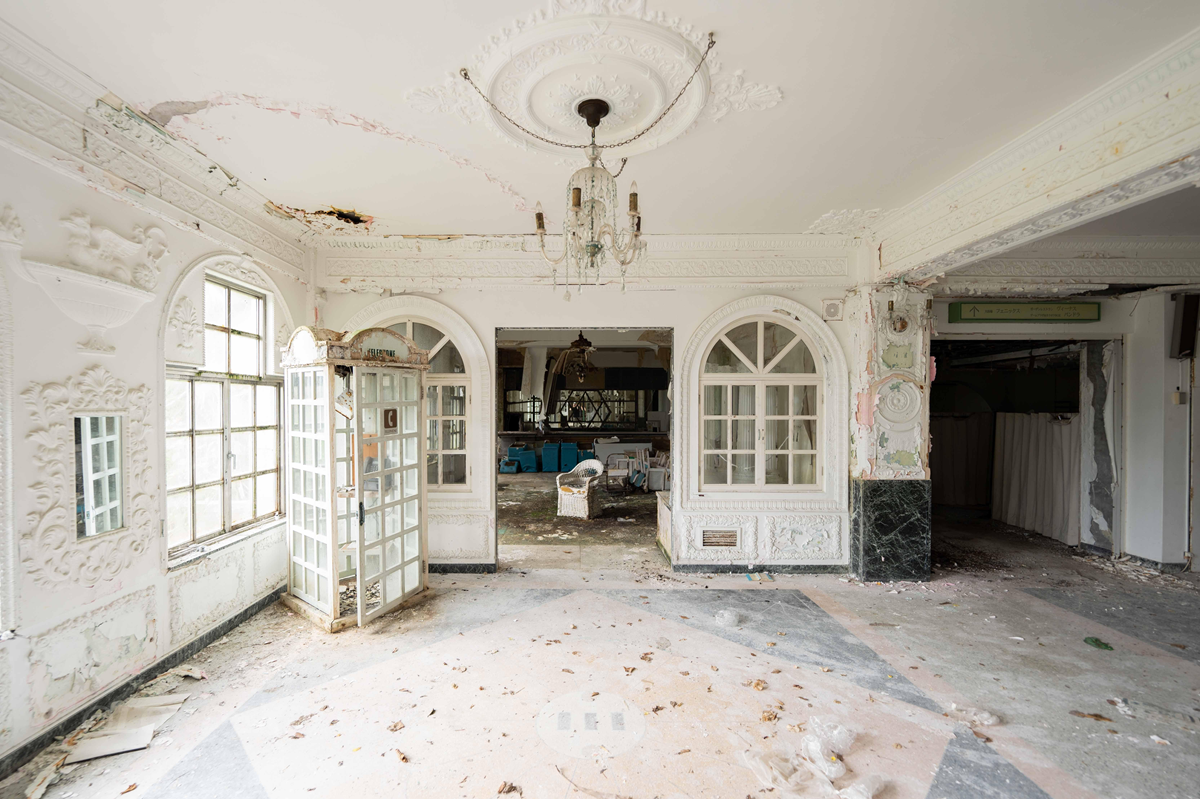 An ornate, abandoned room with peeling paint, a chandelier, arched windows, and a partially open French door. The floor is dirty and debris is scattered. The back area reveals an empty, neglected interior. 6. Hachijo Royal Hotel, Hachijō-jima, Japan