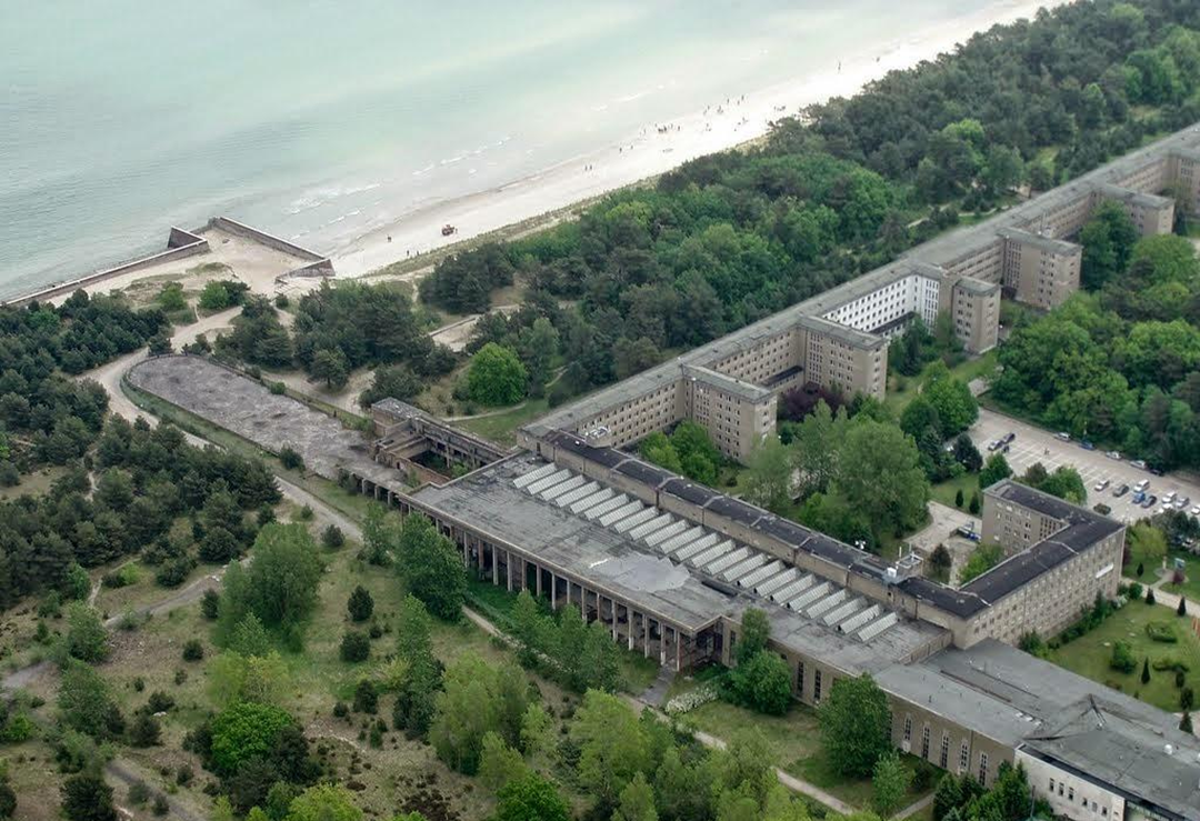 Aerial view of a large, abandoned concrete building complex near a forested coastline and sandy beach, with several rectangular structures surrounded by trees. 13. Prora, Rügen, Germany