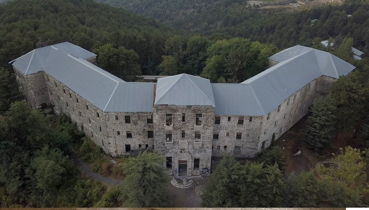 Aerial view of a large, old stone building with a metal roof, surrounded by dense forest and trees, located in a mountainous area. The structure has multiple windows and an angular, U-shaped layout. 12. Berengaria Hotel, Troodos Mountains, Cyprus