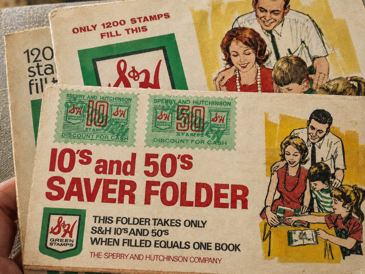 Close-up of vintage S&H Green Stamps saver folders, featuring green stamps and illustrations of smiling families collecting stamps together. Text details the folder’s purpose and the value of the stamps.