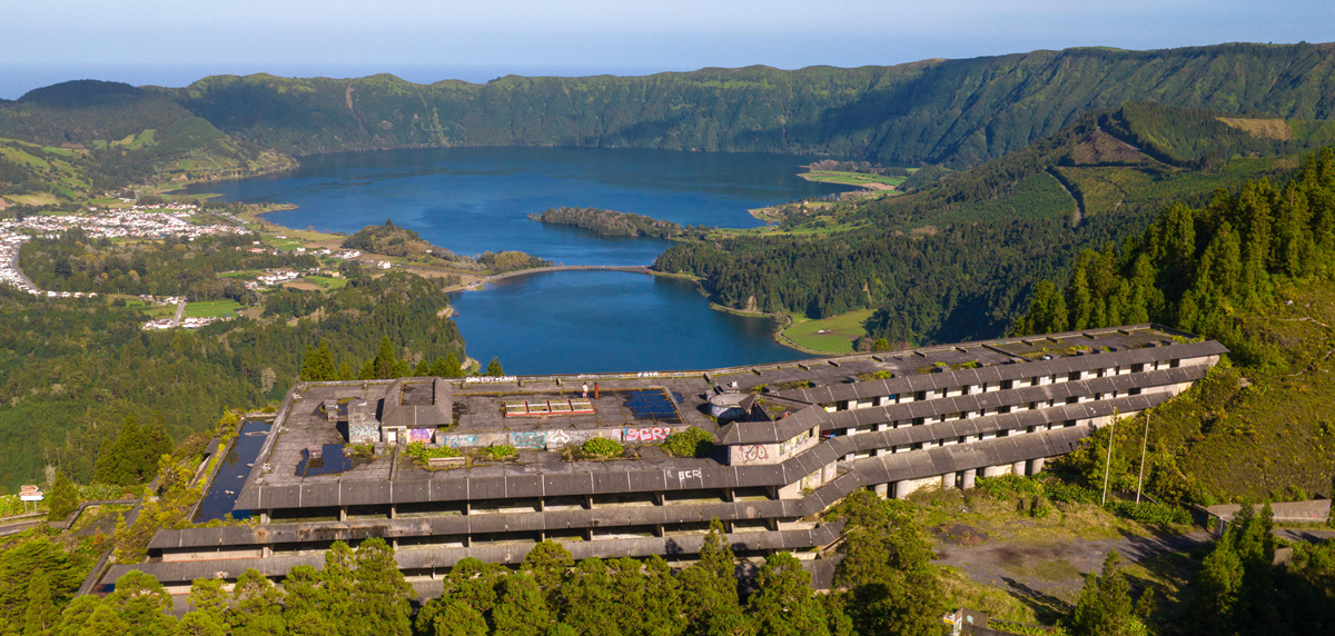 A large, abandoned hotel covered in graffiti sits on a forested hillside overlooking twin blue lakes and a lush volcanic crater, with mountains and a small town in the distance under a clear sky. 2. Monte Palace Hotel, São Miguel, Azores, Portugal