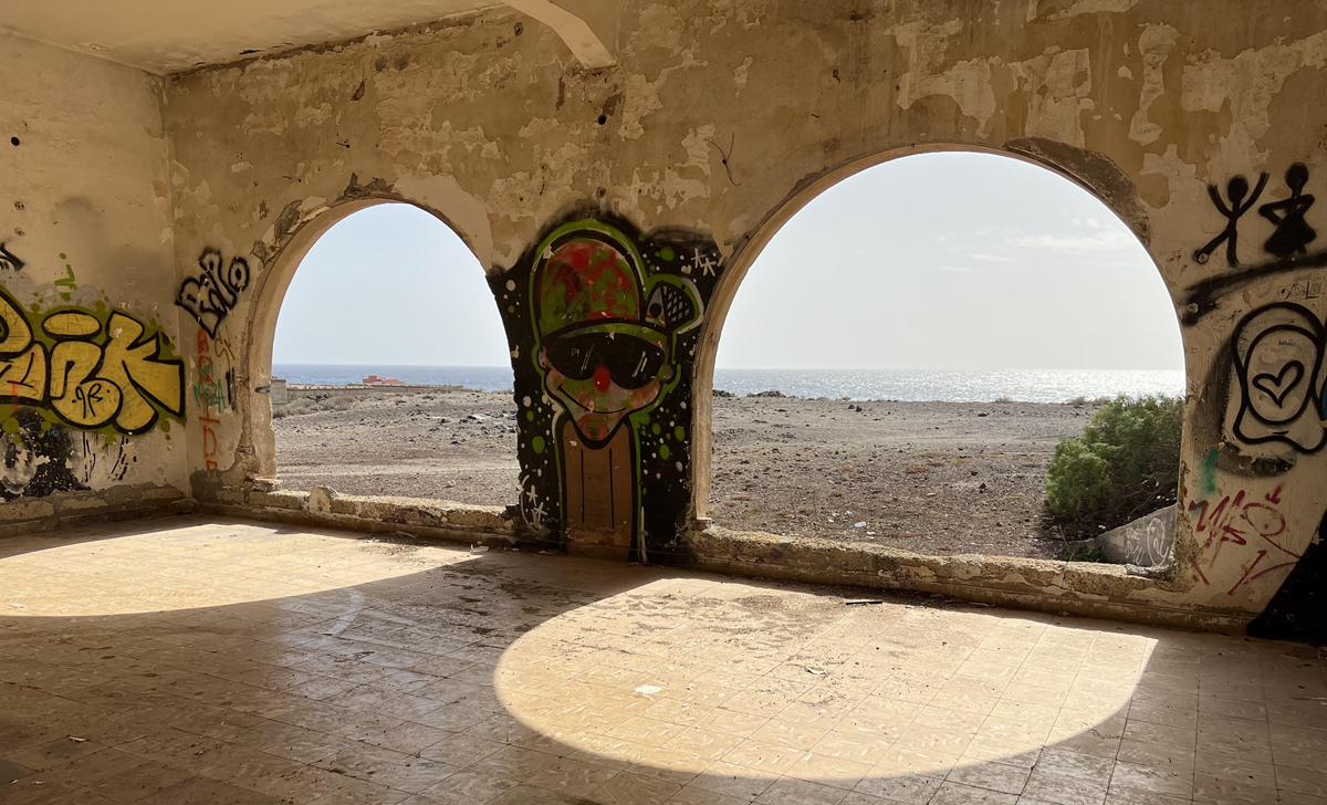 Sunlight streams through three large arched windows in a weathered, graffiti-covered wall, casting shadows on the tiled floor. Through the arches, a rocky beach and the ocean are visible under a bright sky. 11. Sanatorio de Abona, Tenerife, Spain