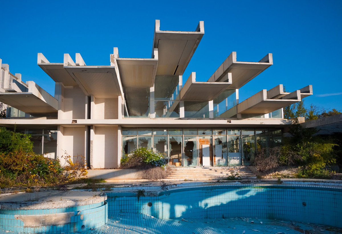 A modernist, abandoned building with exposed concrete beams and large glass windows stands behind an empty, dilapidated swimming pool surrounded by overgrown vegetation under a clear blue sky. 3. Haludovo Palace Hotel, Krk, Croatia