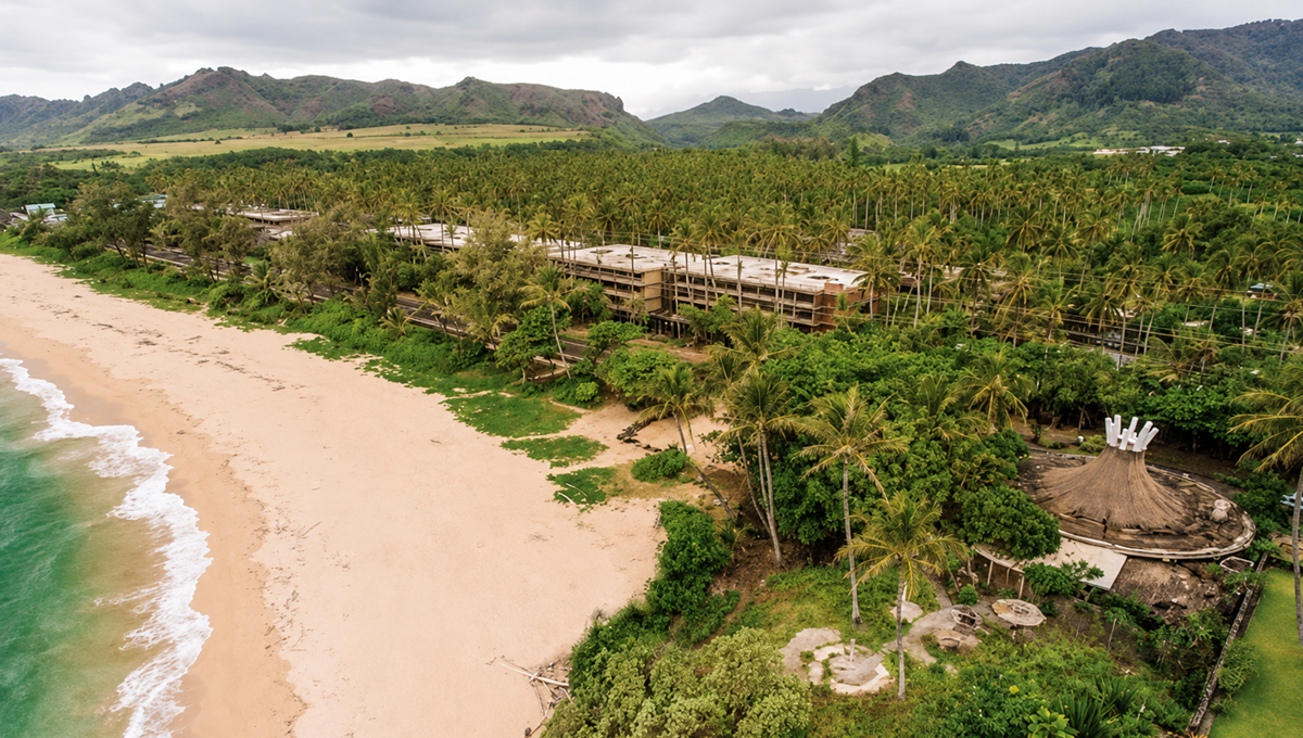 Aerial view of a deserted sandy beach with turquoise water, lush green palm trees, an abandoned resort, and distant mountains under a cloudy sky. 14. Coco Palms Resort, Kauai, Hawaii