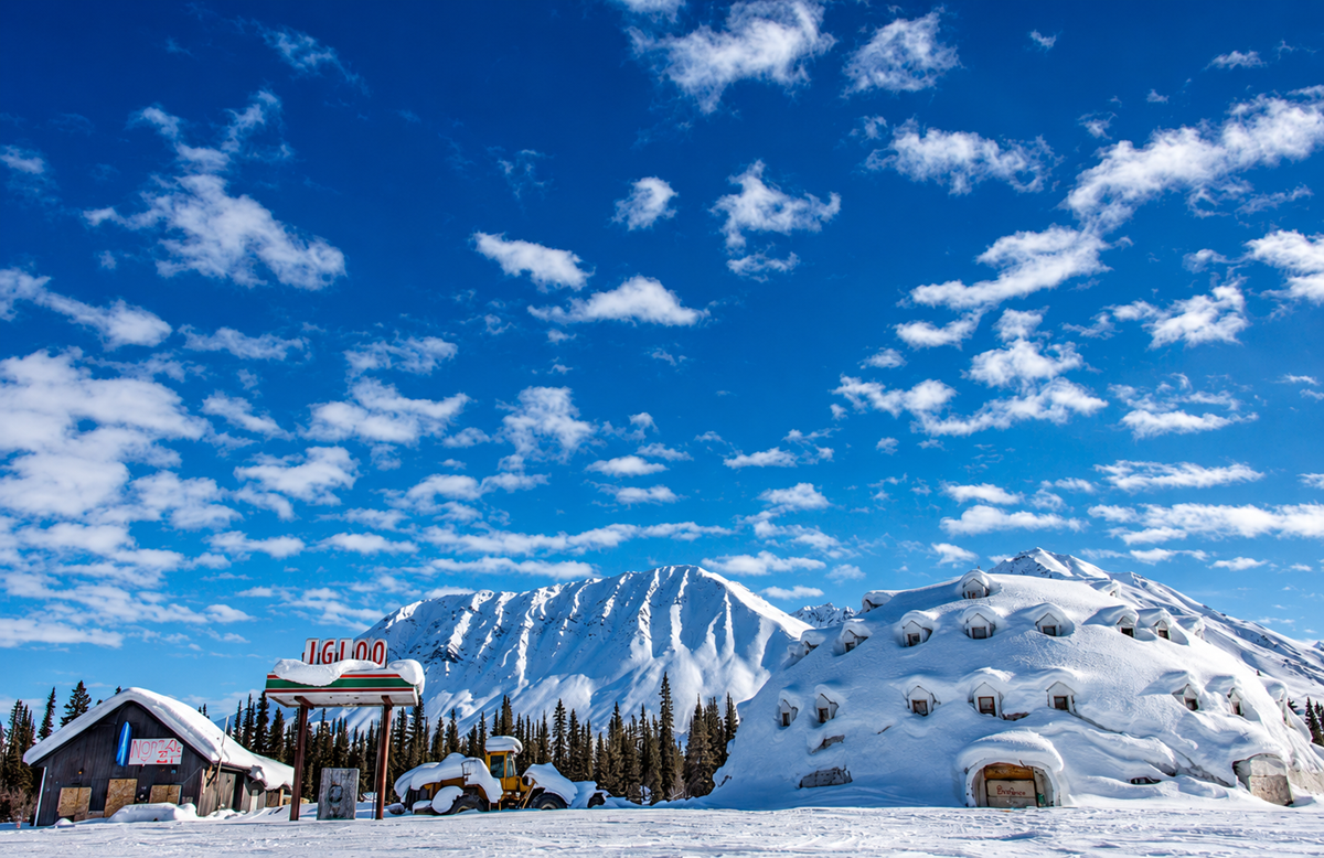 A large igloo-shaped building and a small cabin sit in a snowy landscape with pine trees, backed by tall, snow-covered mountains under a blue sky with scattered clouds. 7. Igloo City, Alaska, United States