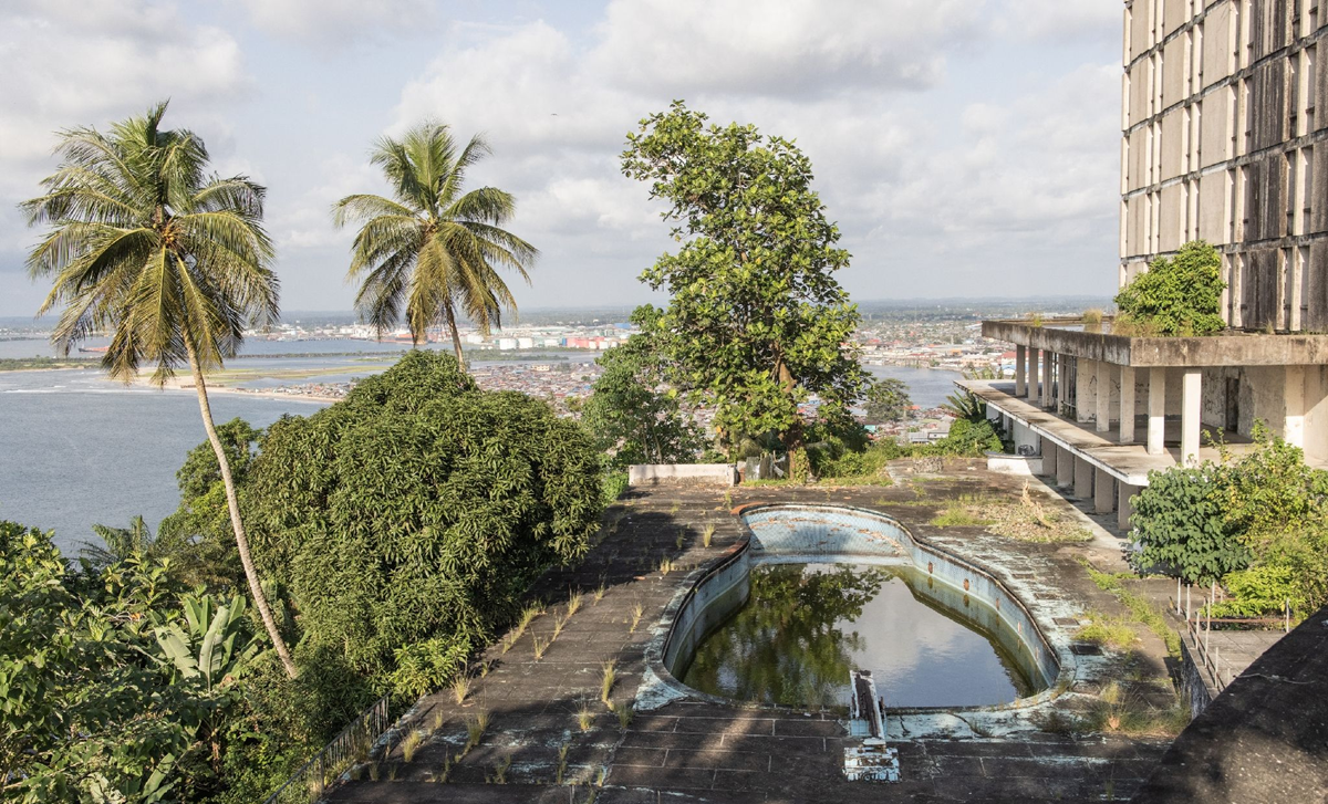 An abandoned rooftop pool with stagnant water and overgrown plants overlooks a coastal cityscape, with palm trees and a large building nearby under a partly cloudy sky. 8. Ducor Hotel, Monrovia, Liberia
