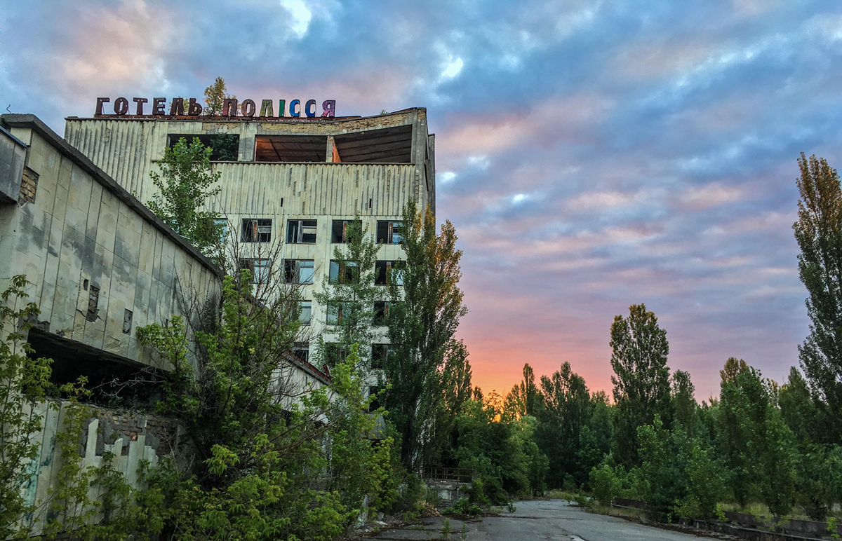 An abandoned, overgrown hotel with a faded sign in Cyrillic script stands under a colorful, cloudy sunset sky, surrounded by tall trees and cracked pavement. 9. Hotel Polissya, Pripyat, Ukraine