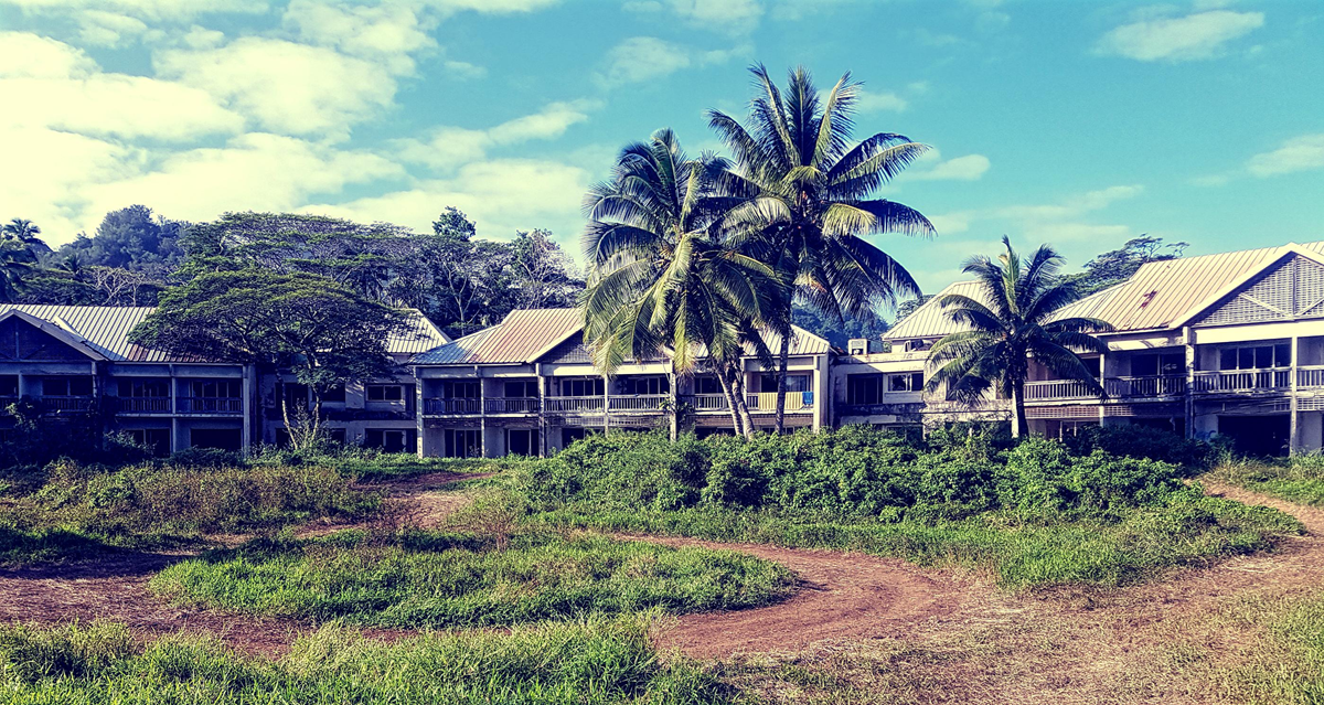 Several unfinished or abandoned buildings stand behind tall grass, palm trees, and other greenery under a bright blue sky with scattered clouds. 10. Sheraton Rarotonga, Cook Islands