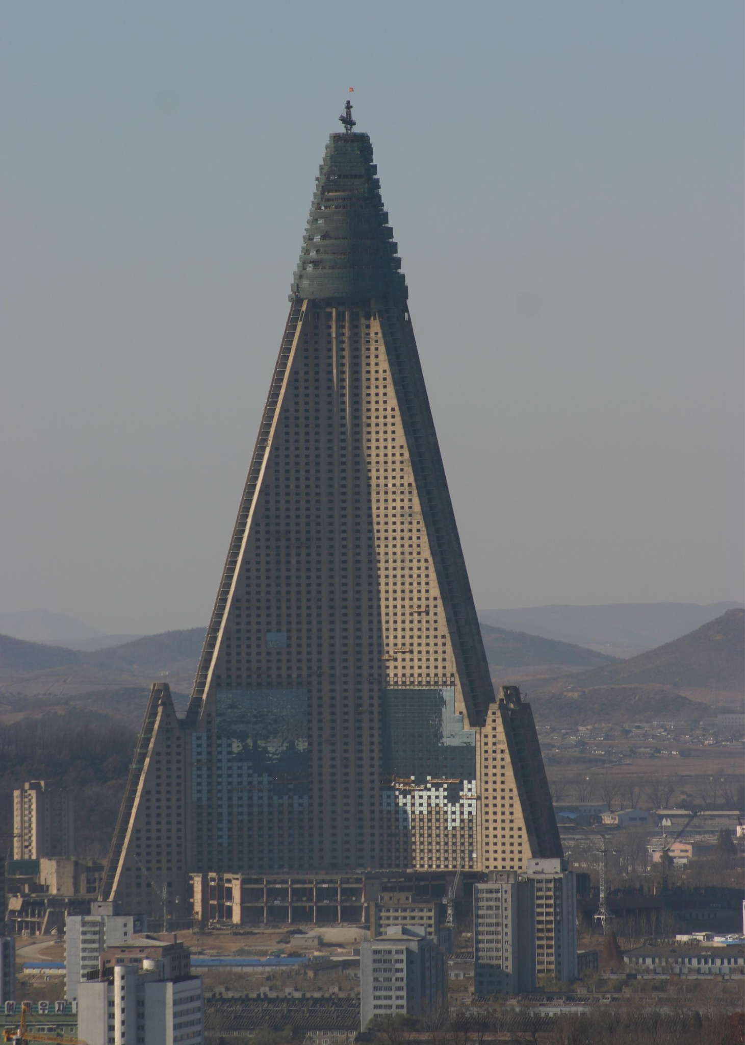 A tall, pyramid-shaped skyscraper with a glass facade and an unfinished appearance stands in a cityscape with smaller buildings and distant mountains in the background under a clear sky. 16. Ryugyong Hotel, Pyongyang, North Korea