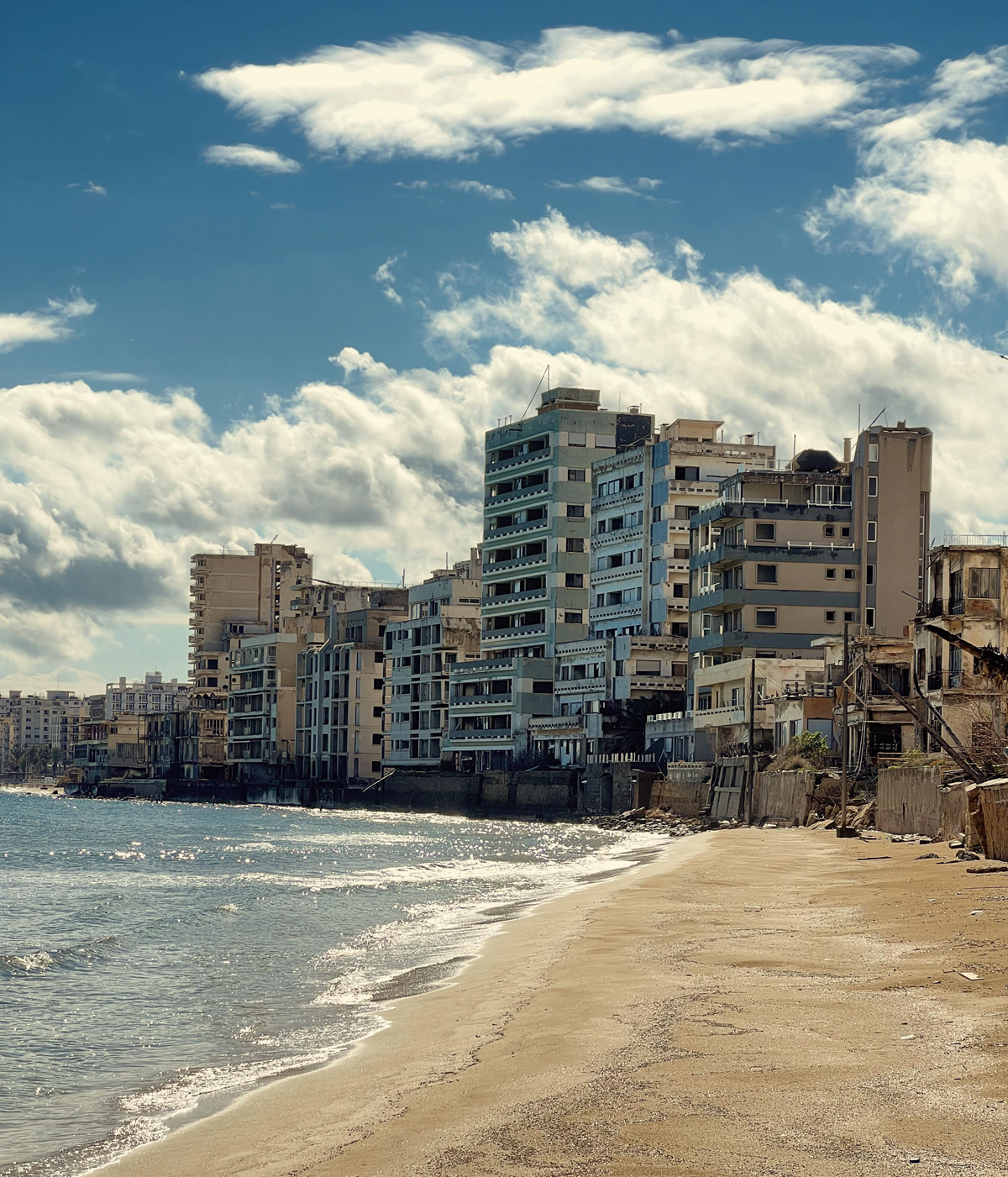 A sandy beach with gentle waves in front of abandoned, weathered high-rise buildings under a partly cloudy blue sky. The scene appears quiet and deserted. 5. Varosha District, Famagusta, Cyprus