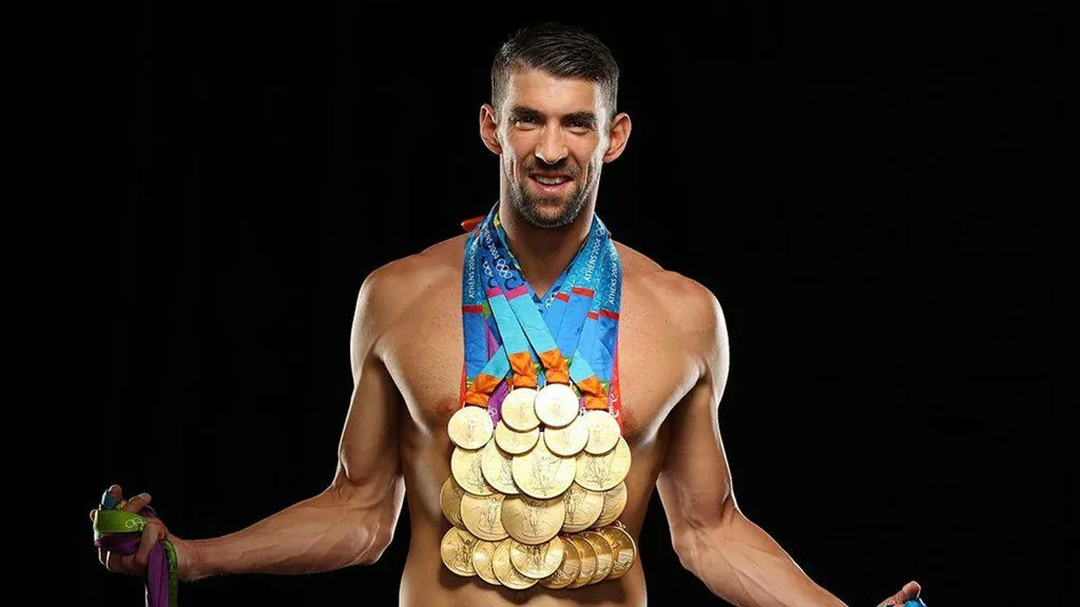 A shirtless male athlete stands smiling, wearing many gold medals around his neck and holding more medals in both hands, against a black background. 7. Michael Phelps’ 23 Olympic Gold Medals
