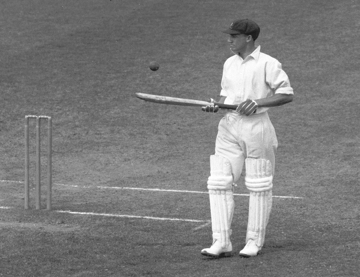 A cricketer in white uniform and pads stands at the crease, holding a bat and watching a cricket ball mid-air. Stumps are behind him on a grassy field. 6. Don Bradman’s 99.94 Test Batting Average