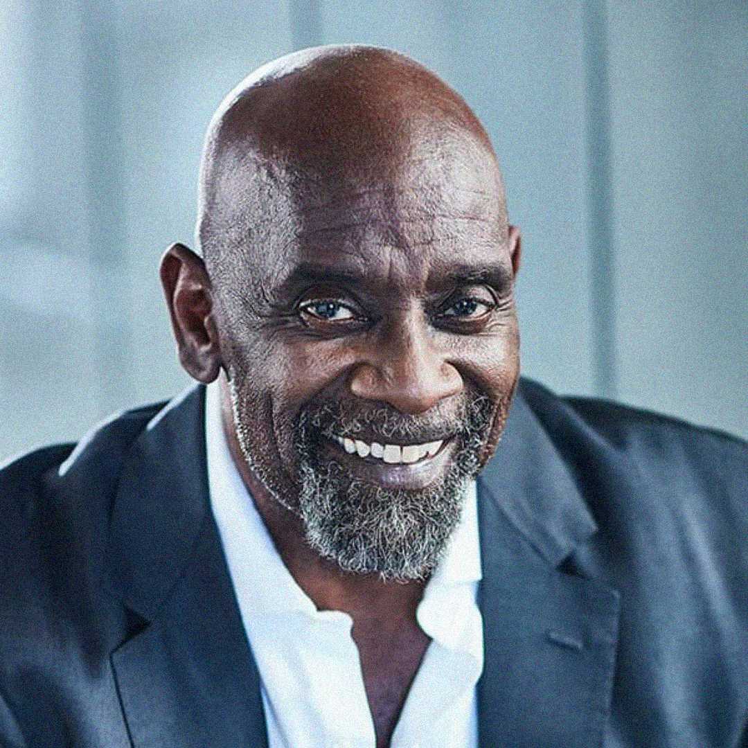 A middle-aged man with a bald head, gray beard, and mustache smiles warmly while wearing a dark blazer and white shirt, sitting in front of a blurred light-colored background.