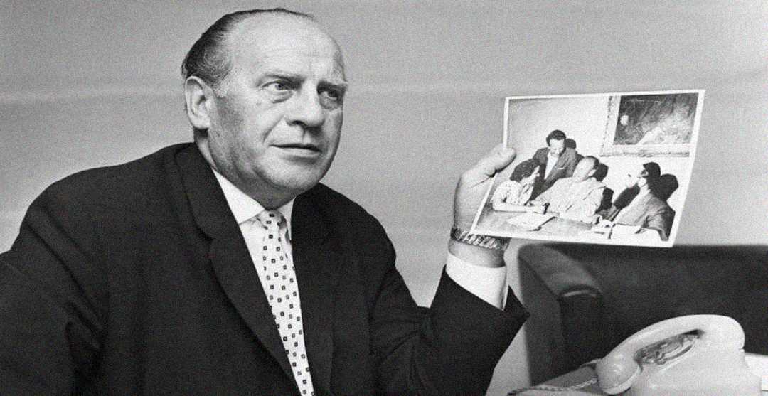 A man in a suit and polka-dot tie sits at a desk, holding up a photograph showing several people seated around a table. An old-fashioned telephone is visible in the foreground.