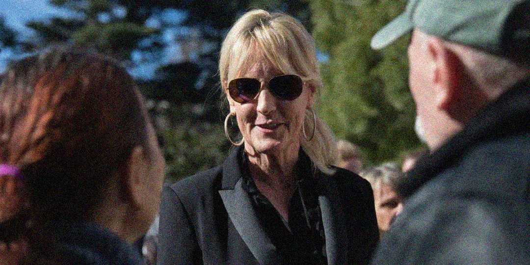 A woman wearing sunglasses, large hoop earrings, and a black blazer is speaking with two people outdoors on a sunny day, with trees in the background.