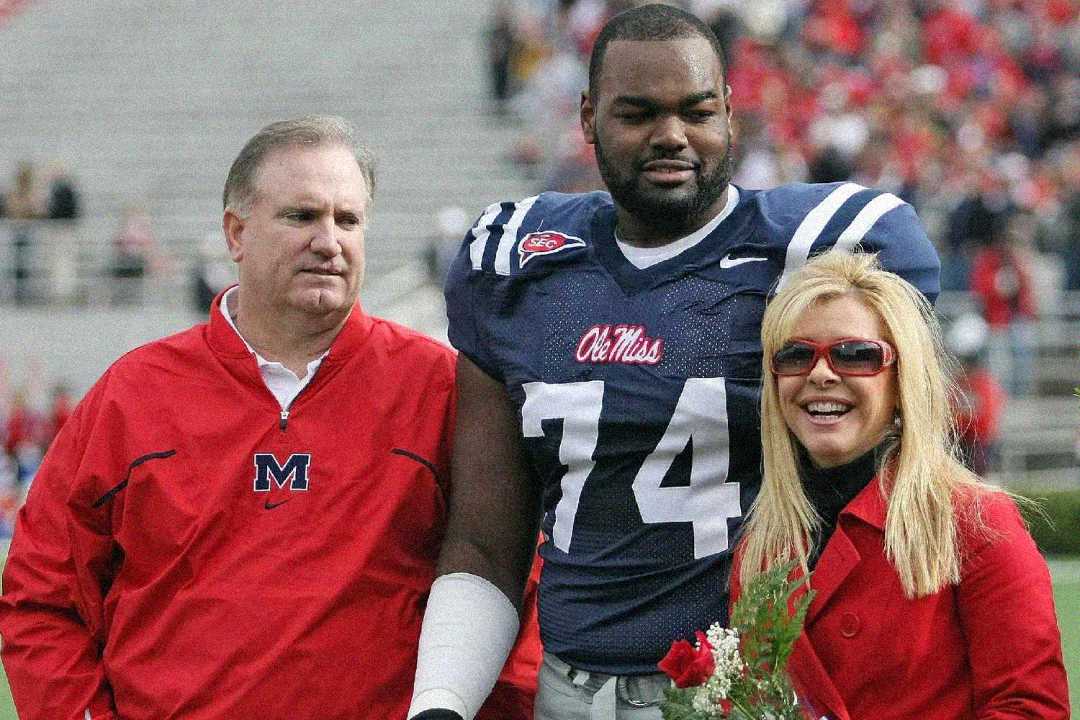 A college football player in an Ole Miss jersey stands on the field with his arms around a man and a woman, both wearing red. The woman holds a bouquet of flowers, and the stadium seats are filled in the background.