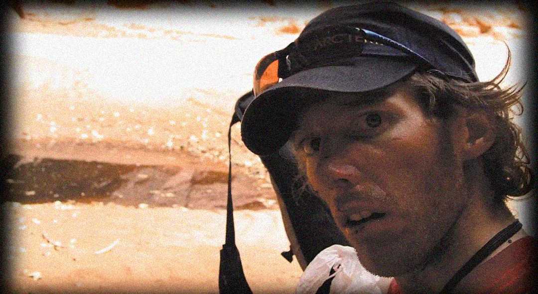 A person with a backpack, wearing a cap and sunglasses on their head, looks wide-eyed at the camera in an outdoor, sandy environment.