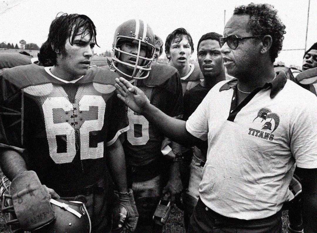 A coach in a "Titans" shirt speaks to a group of American football players in uniform, including player number 62, during what appears to be a practice or game. The scene is intense and focused.