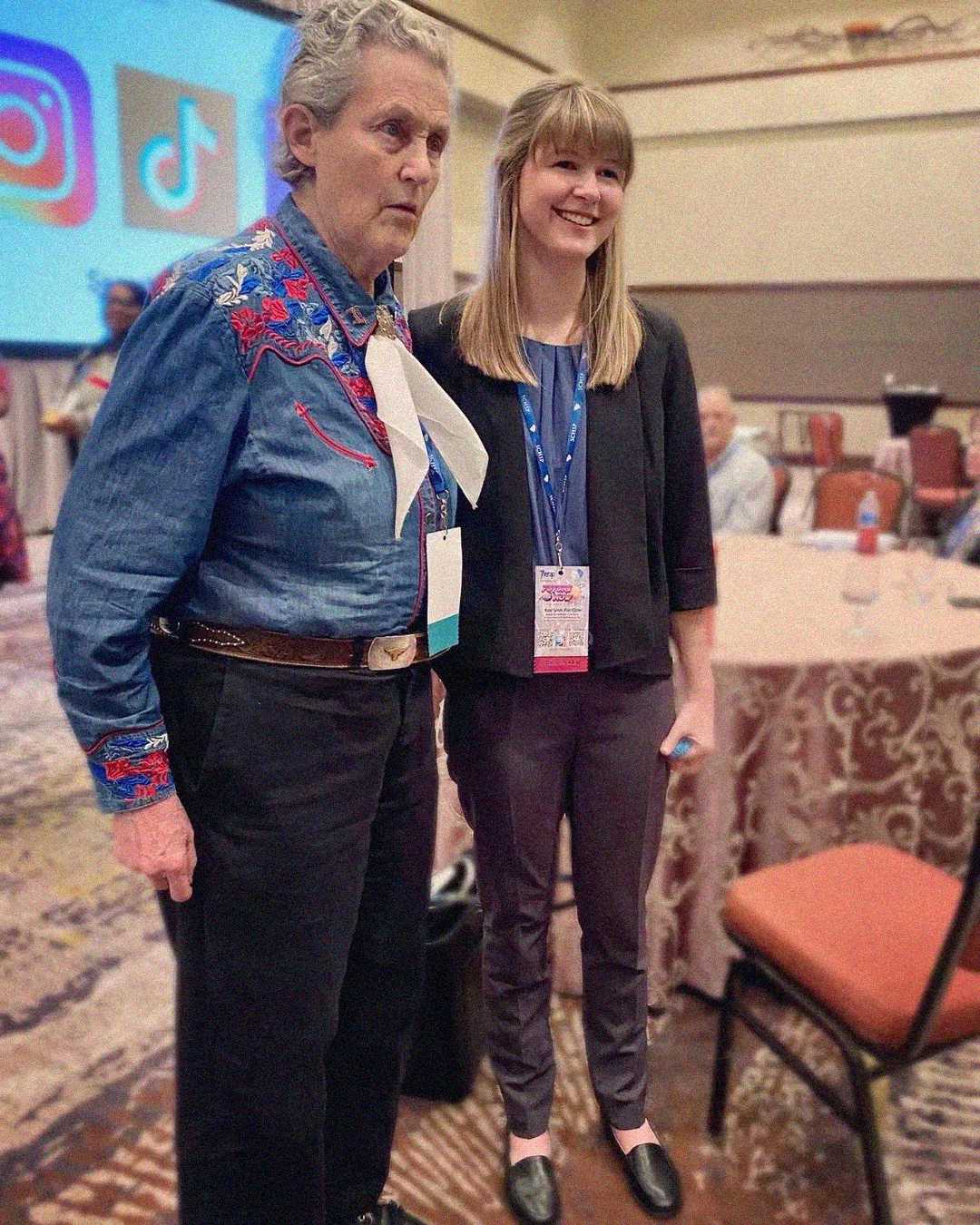 Two women stand together at an indoor event. One wears a blue western-style shirt with red embroidery and a large white scarf; the other wears business casual clothing with a conference badge. People and round tables are in the background.