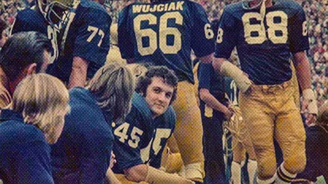 A group of American football players in blue and yellow uniforms sit and stand on the sidelines. Numbers and names are visible on their jerseys, including 45, 66, 68, and 71. One player is looking toward the camera.