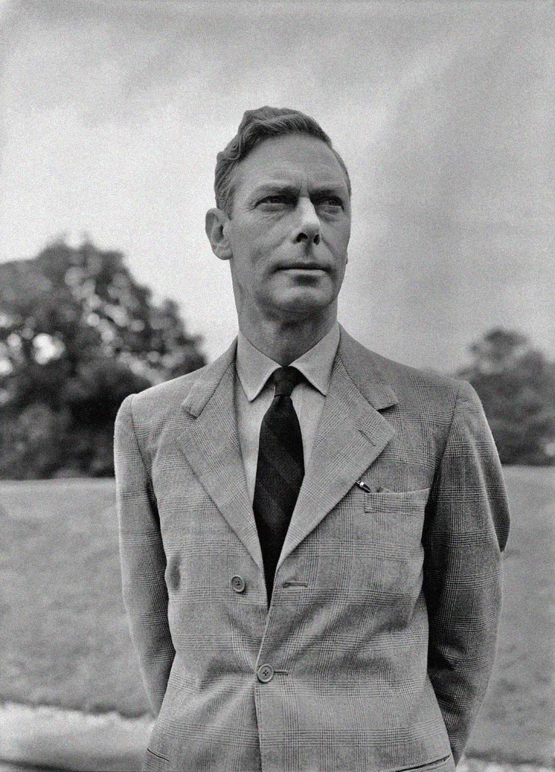 A black-and-white photo of a man in a suit and tie standing outdoors with trees and grass in the background. He is looking slightly to his left with a serious expression and his hands behind his back.