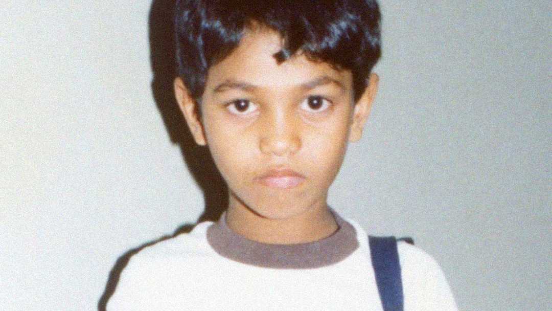 A young boy with short dark hair and a serious expression stands in front of a plain light-colored background, wearing a white shirt with brown trim and a dark strap over his shoulder.