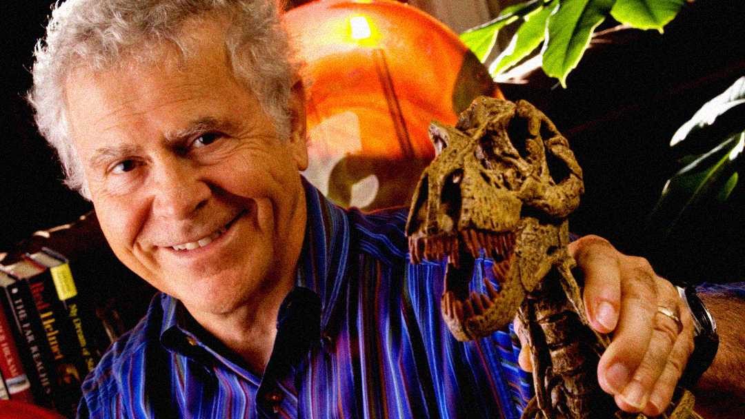 An older man with curly gray hair smiles while holding a small dinosaur skull model. In the background, there are books, a plant, and an orange globe.