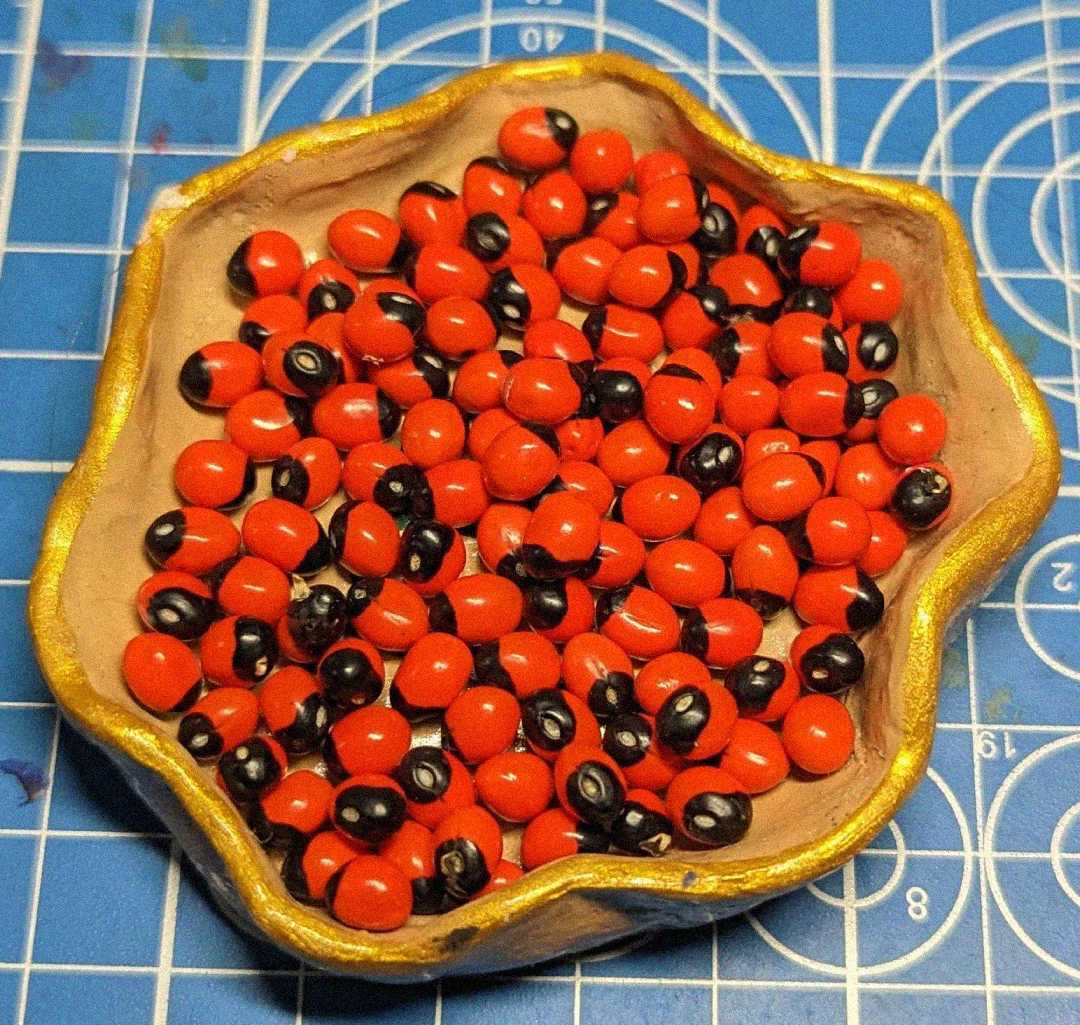 A small, scalloped-edged dish with a gold rim holds numerous bright red, oval seeds, each with a black spot, resembling rosary pea seeds. The dish is placed on a blue grid-patterned background.