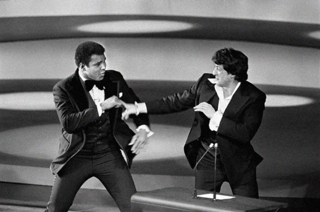 Two men in suits playfully pretend to box on stage, smiling and facing each other under bright spotlights, with a microphone stand visible in front of them.