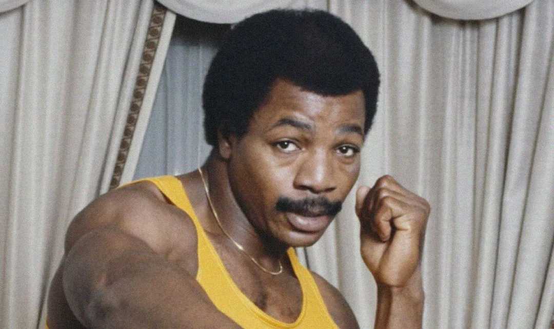 A man with a mustache wearing a yellow tank top poses with his fists raised in a boxing stance, standing in front of cream-colored curtains.