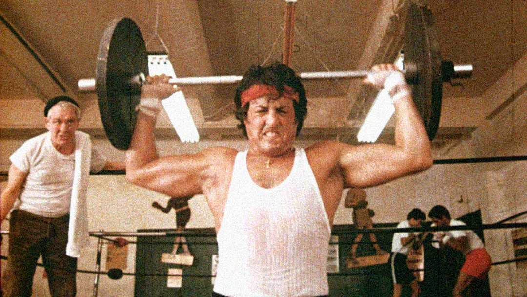 A man wearing a white tank top and red headband lifts a barbell over his head with a strained expression in a gym, while people exercise in the background.