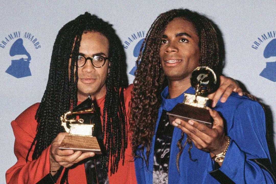 Two men with long braided hair hold Grammy trophies while posing together in front of a backdrop with the Grammy Awards logo. One wears glasses and a red jacket; the other wears a blue jacket and smiles.