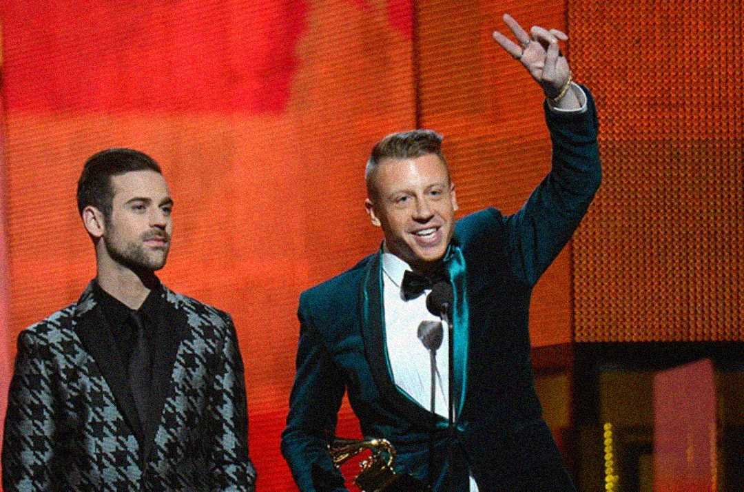 Two men in suits stand on stage at an awards show. One man holds a trophy and raises his hand in a peace sign, smiling, while the other stands beside him looking ahead. A microphone and red backdrop are visible.