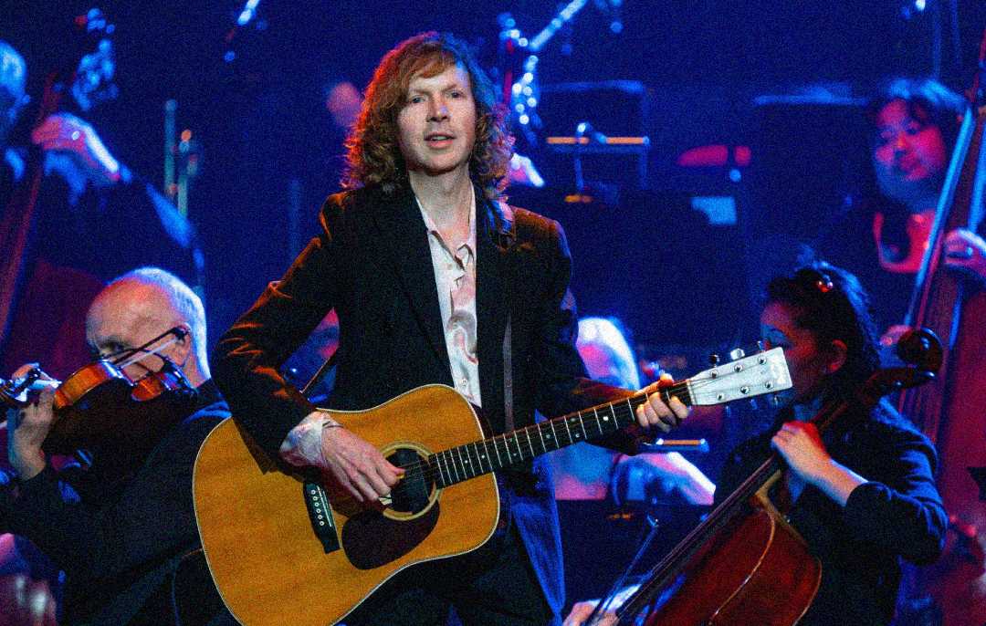A musician with wavy, shoulder-length hair plays an acoustic guitar on stage, surrounded by an orchestra with string instruments, under blue and purple stage lighting.
