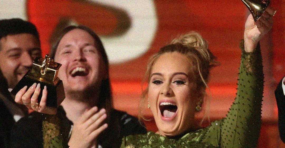 A woman in a green dress joyfully raises a Grammy award while people around her smile and applaud, celebrating on stage.