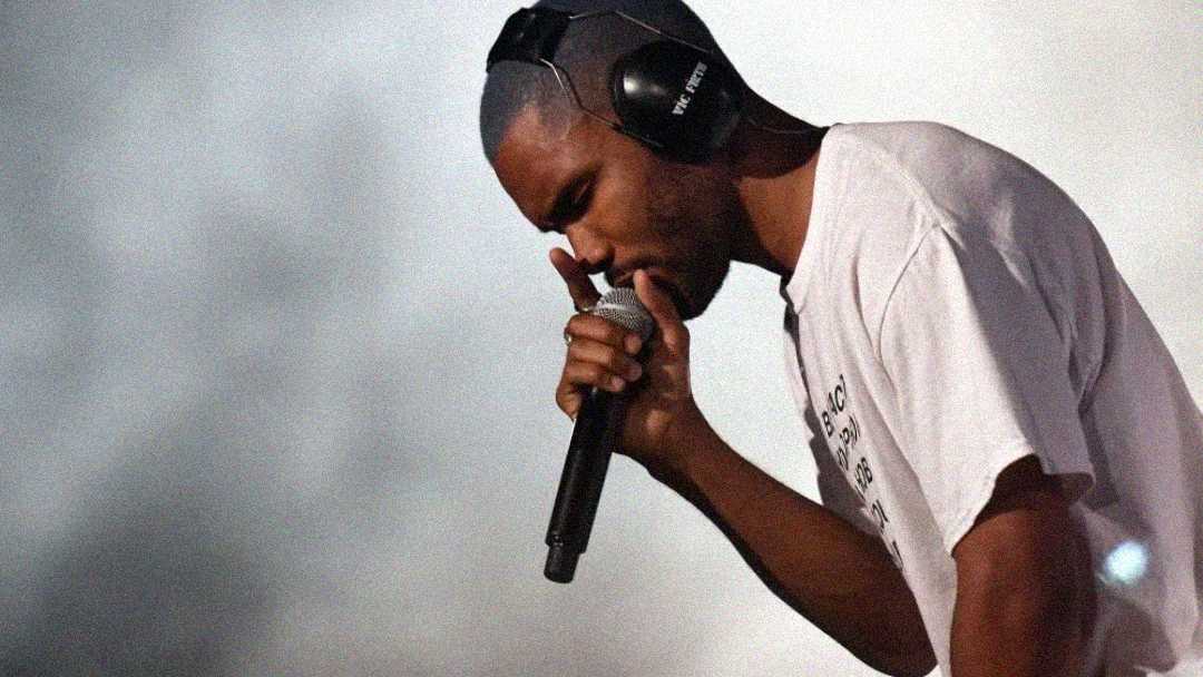 A man in a white t-shirt and black headphones holds a microphone close to his mouth, appearing to sing or speak, with a blurred light background behind him.