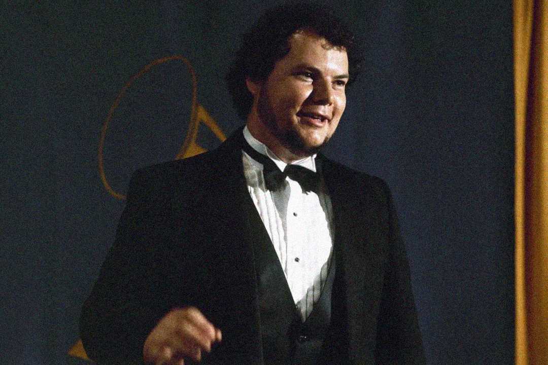 A man in a black tuxedo and bow tie stands in front of a dark backdrop with a faint gold outline, possibly an award symbol, smiling and gesturing with one hand.