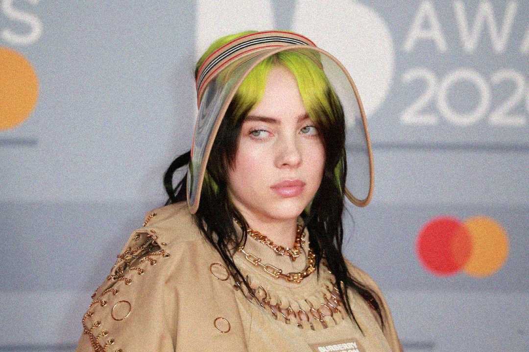 A person with black and neon green hair wears a tan outfit with chains and a wide-brimmed hat with ribbons, standing in front of a blurred background featuring the text "AWARDS 2020" and colorful circles.