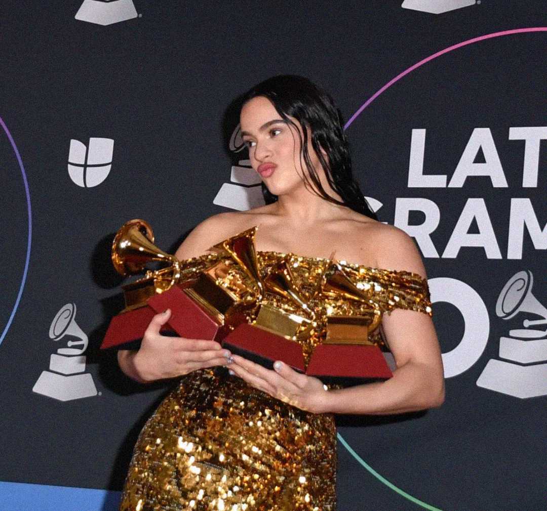 A woman in a gold sequin dress holds three Grammy awards and poses in front of a backdrop featuring the Latin Grammy Awards logo.