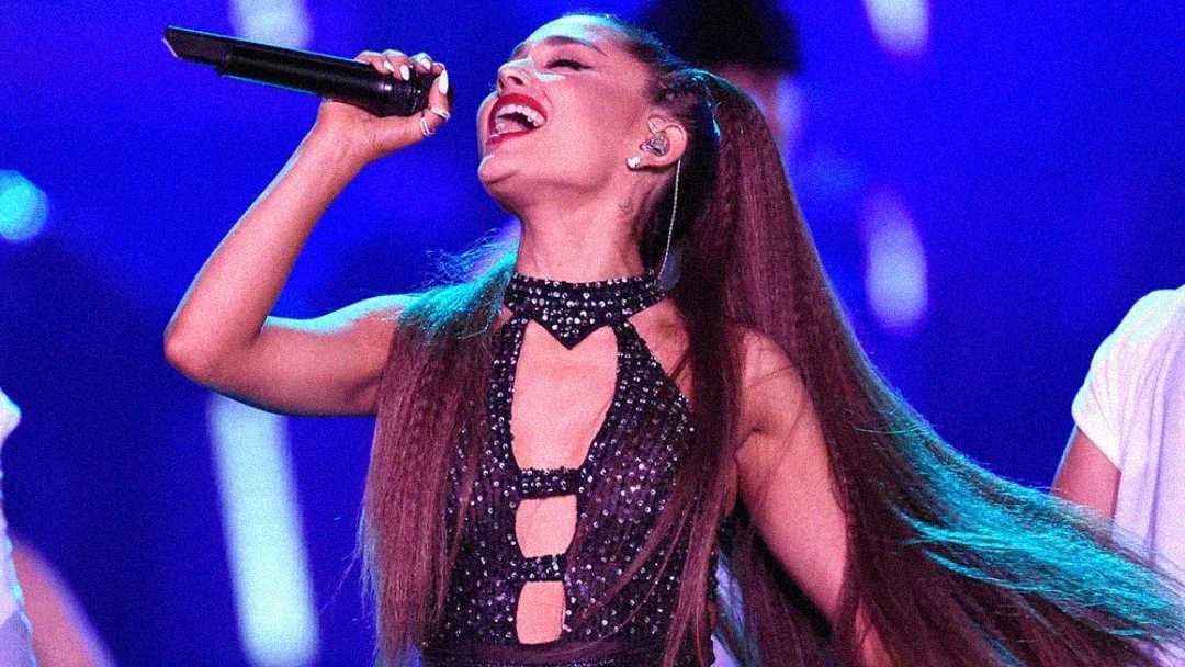 A woman with long brown hair in a high ponytail sings passionately into a microphone on stage, wearing a sparkly black outfit against a vibrant blue and purple background.