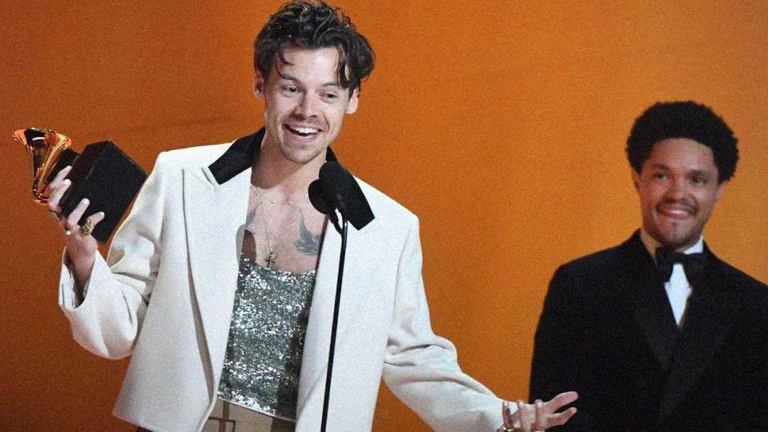 A smiling man in a white jacket holds up a trophy while speaking at a microphone on stage, with another man in a black suit standing and smiling in the background against an orange backdrop.