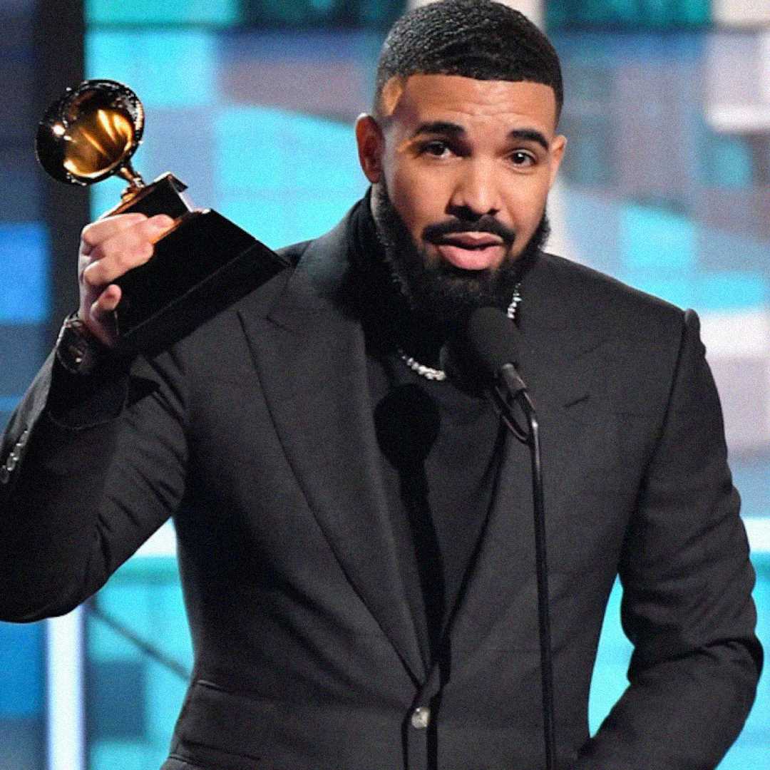 A man with a trimmed beard and short hair, dressed in a dark suit, holds up a Grammy award trophy while speaking at a microphone on stage.
