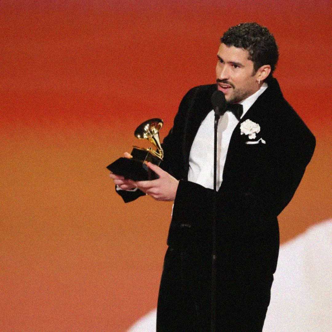 A man in a black tuxedo holds a Grammy award and speaks into a microphone on stage, with a warm orange background behind him.