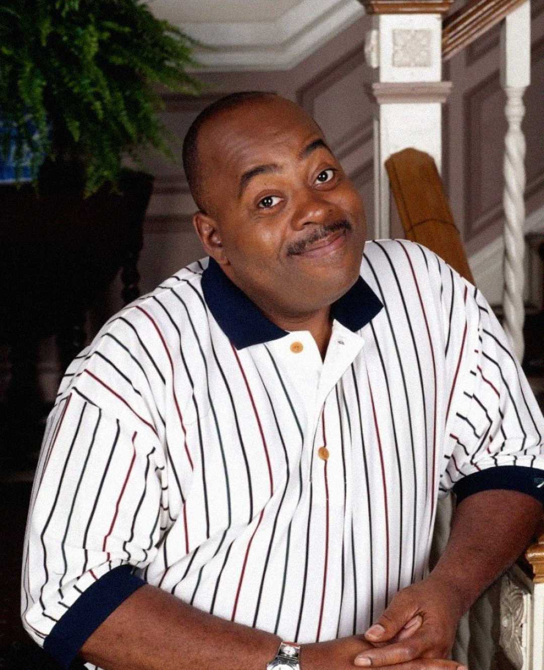 A smiling man with a mustache wearing a white polo shirt with black and red stripes stands near a staircase inside a house.
