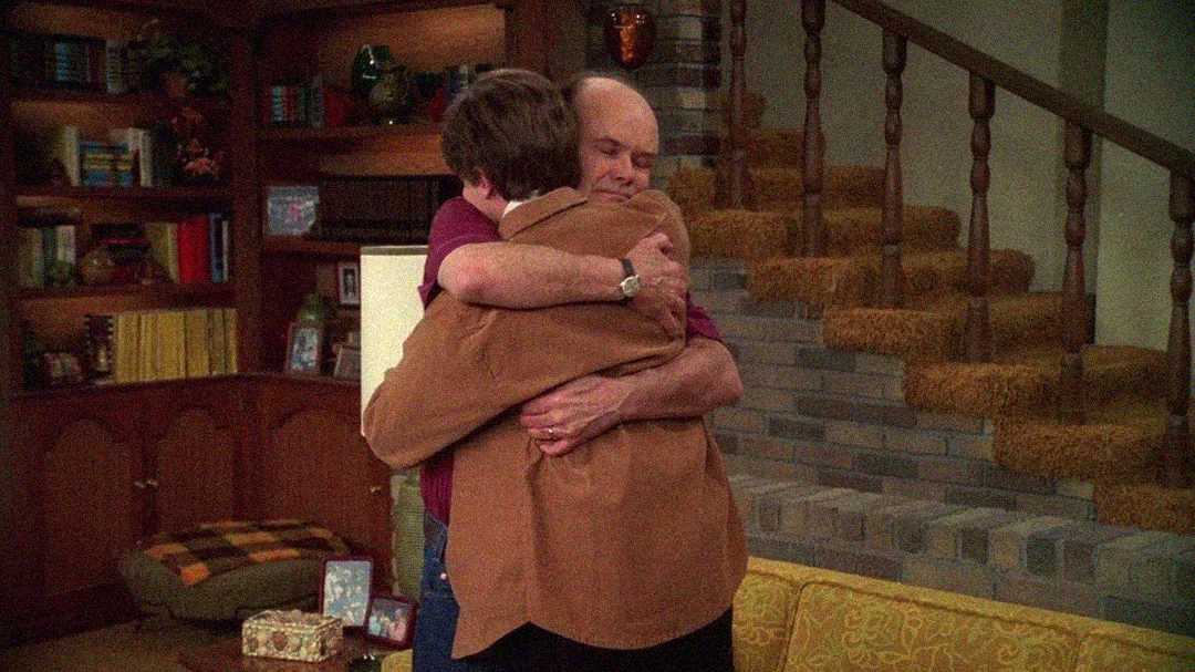 Two people share a heartfelt hug in a cozy living room, with shelves, a brick staircase, and a yellow couch in the background. One person wears a brown jacket, the other a red shirt.