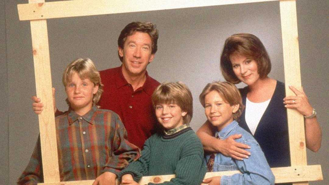 A family of five poses together, smiling, while holding a large wooden frame. The adults stand behind three boys, all dressed in casual 90s-style clothing, against a plain background.