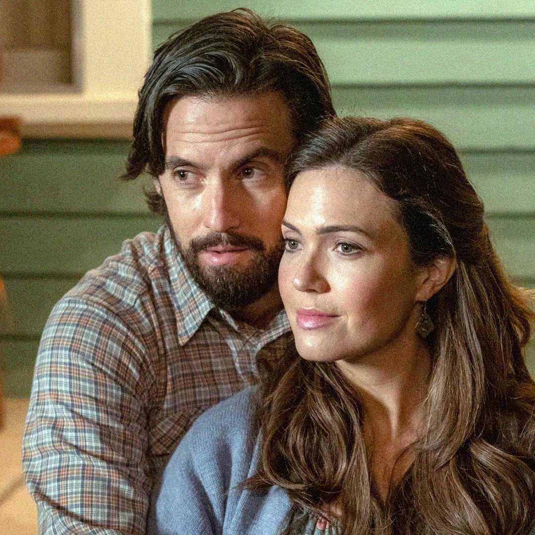 A man with dark hair and a beard hugs a woman with long brown hair. They both look thoughtfully into the distance, standing in front of a green wooden wall. The mood appears calm and affectionate.