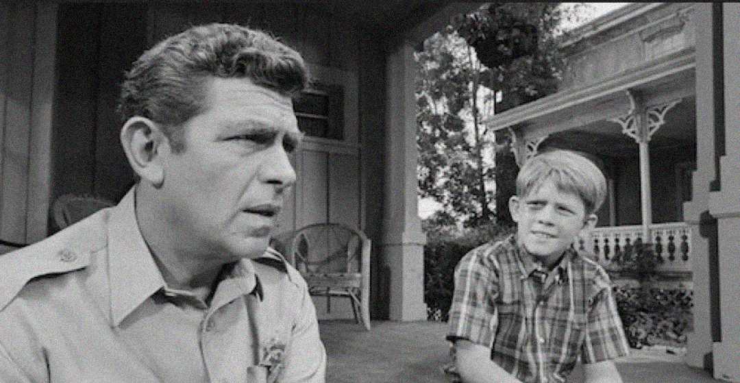 A black-and-white photo of a man in uniform sitting on a porch next to a young boy in a plaid shirt. Both look thoughtful, with a wooden house and trees in the background.