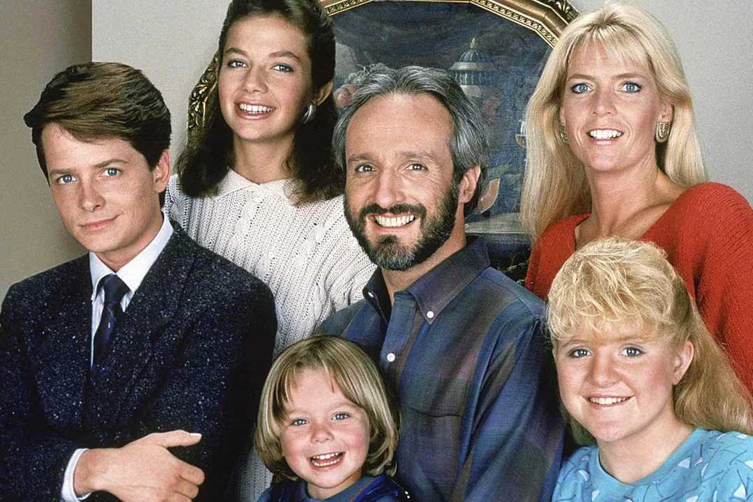A group of six people, including two men, three women, and one young girl, smile and pose together indoors in front of a framed painting. They appear to be a family.