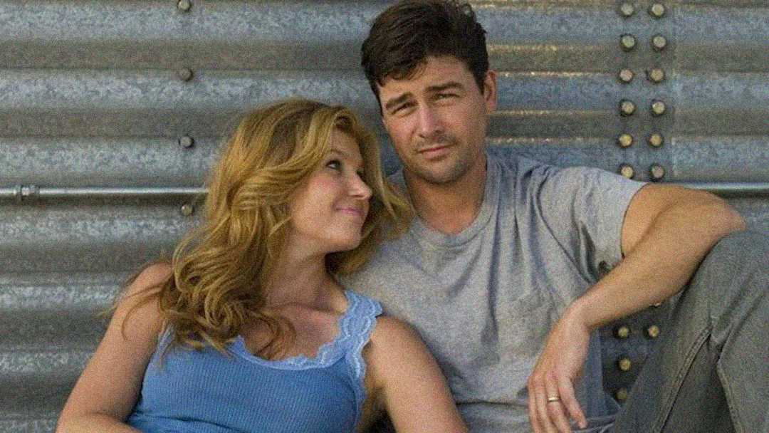 A woman with wavy blonde hair in a blue tank top leans affectionately on a man in a gray t-shirt. They sit together against a corrugated metal wall, looking relaxed and content.